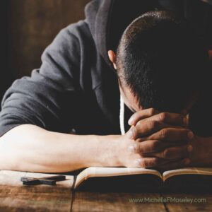 Individual with short hair and wearing a black hoodie has his head down over the Bible and appears in distress.