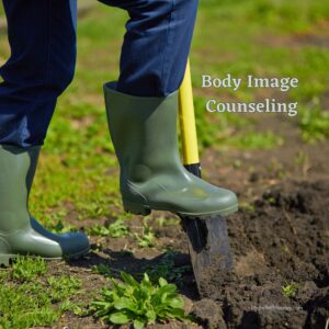 A foot in a dark green waterproof boot pushing down on a shovel to symbolize the work of digging into body image concerns.