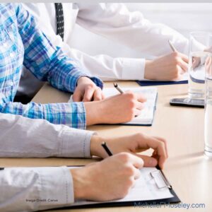 People's hands sorting through papers on a desk. There is also a laptop and a cup of coffee on the desk.