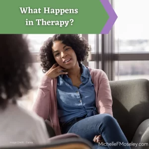 A woman with light brown skin is weating a blue shirt and pink cardigan. She is sitting in a chair facing another person to represent talking to a therapist.