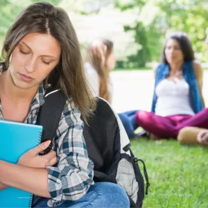 A young white woman with long brown hair is in the forefront of the image. There are other people gathered in a circle behind her. She appears to feel sad and isolated due to being excluded for being different.
