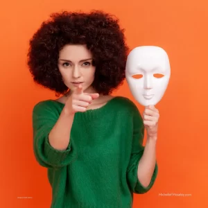 A woman with afro-style curly hair and wearing a green sweater is holding a white theatrical mask that she has pulled away from her face to represent the process of participating in a neurodivergent-affirming assessment.
