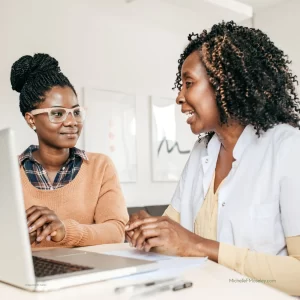 A young Black woman in a peach sweater sits to the left of a slightly older Black woman in a white button-down shirt. They are looking at a computer together as they consult about an important topic.