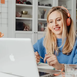 A white woman with long blonde hair and wearing a blue shirt and peach-colored over-ear headphones sits at a computer as she receives specialized consultation on her work.