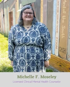 Image of Michelle F. Moseley, Licensed Clinical Mental Health Counselor - a fat, white woman with shoulder length brown-grey hair wearing eyeglasses and a dress a blue pattern.