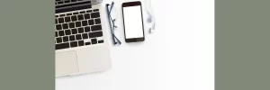 Overhead view of a laptop keyboard, eyeglasses, a cell phone, and ear buds to symbolize preparing technology for a telehealth therapy session.  