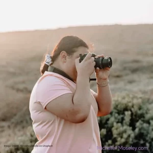 A plus-size person wearing a pale pink shirt is holding a camera up to their face and pointing it toward the horizon to show the freedom found through Body Image Counseling.