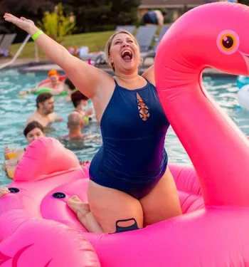 A white woman in a navy blue one-piece swimsuit sits atop a hot pink swan float in a pool. The woman has her arms spread wide and a large smile on her face.