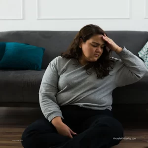 A white woman with long brown hair and weating a grey shirt is sitting on the floor with her head in her hand showing distress related to navigating the world as a neurodivergent person.